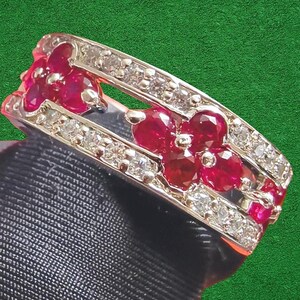 May include: A silver ring with a floral design. The ring features vibrant red ruby gemstones arranged in flower shapes, interspersed with small, sparkling diamonds. The ring is set against a green background.