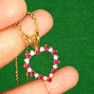 May include: A gold-colored necklace with a heart-shaped pendant. The pendant is adorned with alternating red and white gemstones. The necklace is held against a green background.