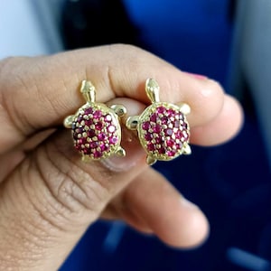 May include: A pair of gold-colored turtle stud earrings. Each turtle has a shell covered in small, round, red gemstones. The earrings are displayed against a blurred background.