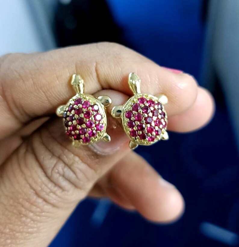 May include: A pair of gold-colored turtle stud earrings. Each turtle has a shell covered in small, round, red gemstones. The earrings are displayed against a blurred background.