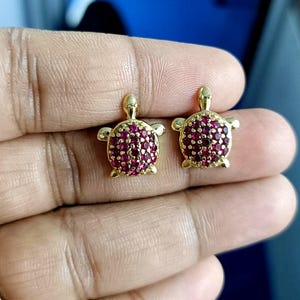 May include: A pair of gold-colored turtle stud earrings. Each turtle has a body covered in small, round, red gemstones. The earrings have a classic post-and-clutch closure.