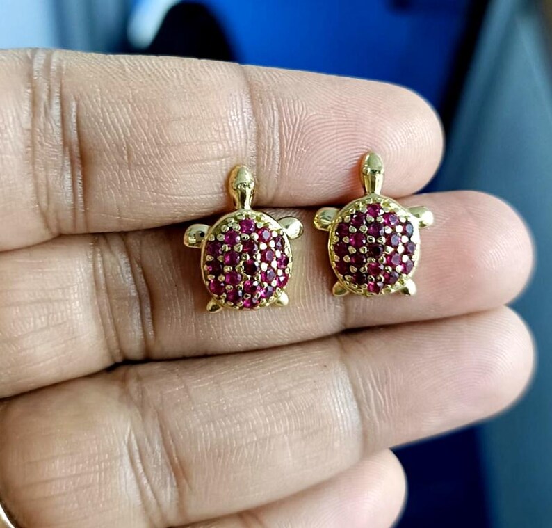 May include: A pair of gold-colored turtle stud earrings. Each turtle has a body covered in small, round, red gemstones. The earrings have a classic post-and-clutch closure.