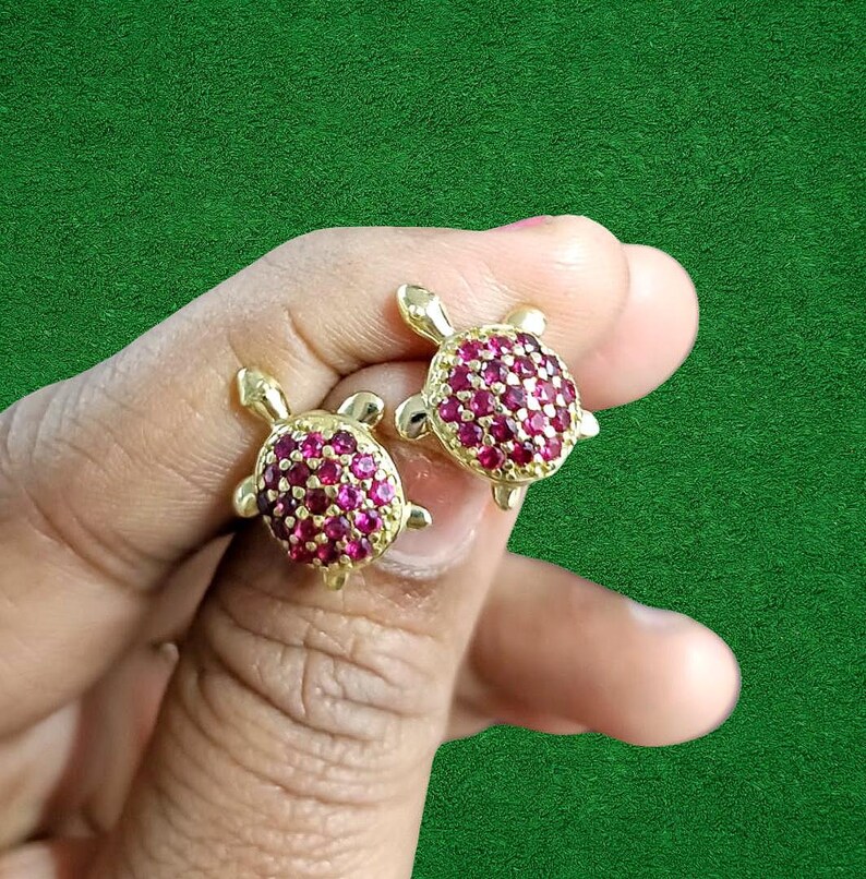 May include: A pair of gold-colored turtle stud earrings. The turtle shells are covered in small, round, ruby-colored stones. The earrings are held against a green background.