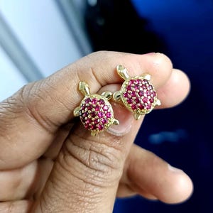 May include: A pair of gold-colored turtle stud earrings. The turtle shells are encrusted with small, round, red gemstones. The earrings are held in a person's hand against a blurred blue background.