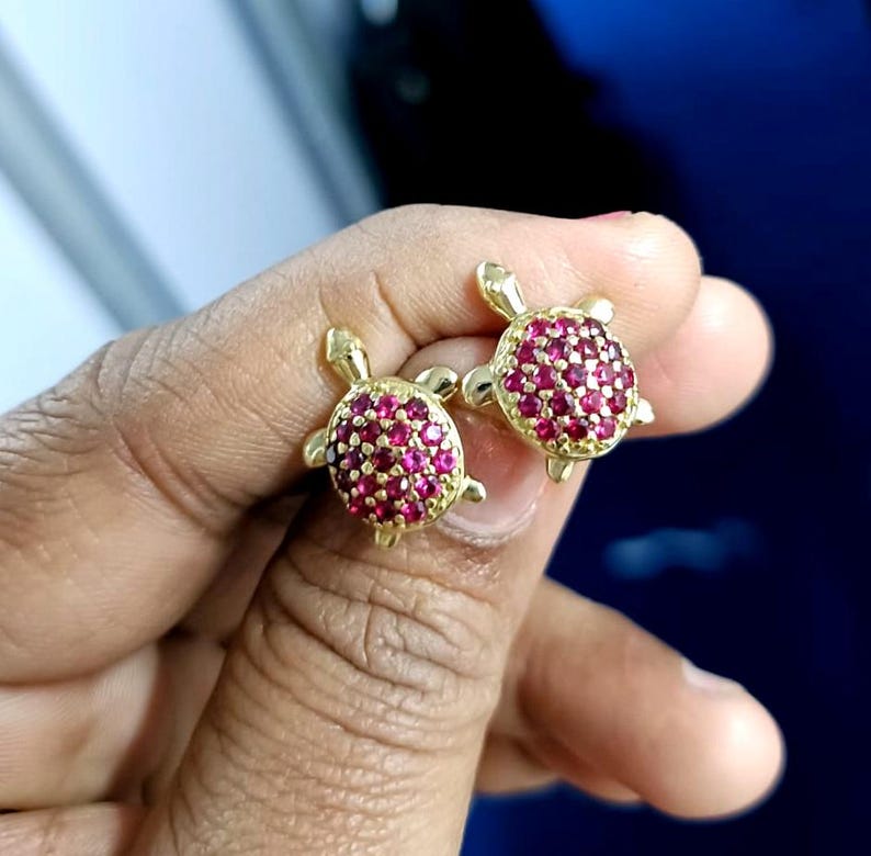 May include: A pair of gold-colored turtle stud earrings. The turtle shells are encrusted with small, round, red gemstones. The earrings are held in a person's hand against a blurred blue background.