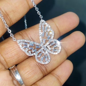 Peut inclure: Un collier pendentif papillon argent&eacute; avec une cha&icirc;ne d&eacute;licate. Le motif papillon est incrust&eacute; de pierres pr&eacute;cieuses claires et scintillantes. Le collier est pr&eacute;sent&eacute; sur une main.