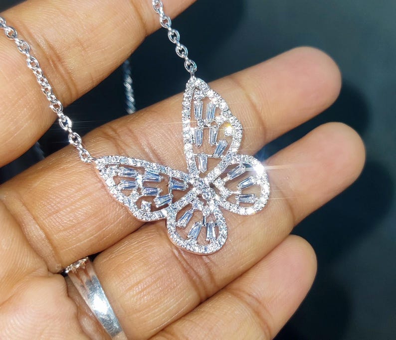 Peut inclure: Un collier pendentif papillon argent&eacute; avec une cha&icirc;ne d&eacute;licate. Le motif papillon est incrust&eacute; de pierres pr&eacute;cieuses claires et scintillantes. Le collier est pr&eacute;sent&eacute; sur une main.