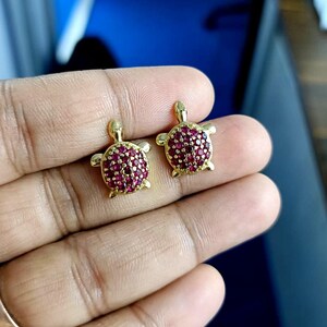 May include: A pair of gold-colored turtle stud earrings. The turtle shells are covered in small, round, red gemstones. The earrings are displayed against a blurred background.
