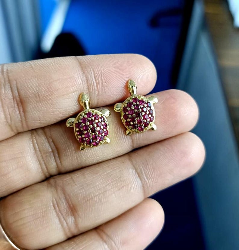 May include: A pair of gold-colored turtle stud earrings. The turtle shells are covered in small, round, red gemstones. The earrings are displayed against a blurred background.