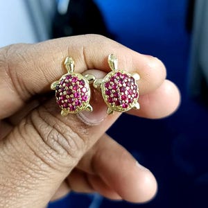 May include: A pair of gold-colored turtle stud earrings. The turtle shells are encrusted with small, round, red gemstones. The earrings are held up against a blue background.
