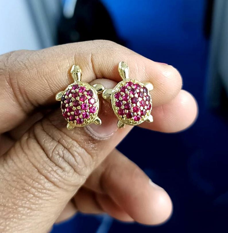 May include: A pair of gold-colored turtle stud earrings. The turtle shells are encrusted with small, round, red gemstones. The earrings are held up against a blue background.