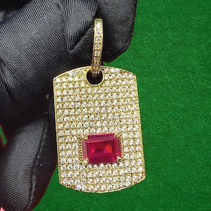 May include: A gold-coloured dog tag pendant, covered in small, clear gemstones. A rectangular, deep red gemstone is set in the centre. The pendant has a loop at the top for a chain. The background is green.