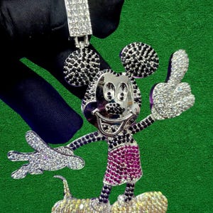 May include: A silver-toned pendant in the shape of Mickey Mouse, encrusted with sparkling crystals and black accents. The pendant features a thumbs-up gesture and a smiling face, with a matching bail at the top for a chain.