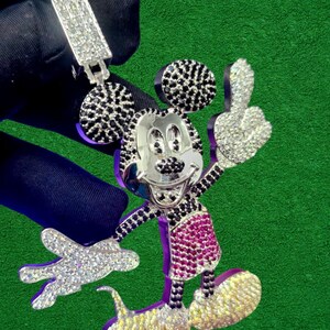 May include: A silver pendant featuring a cartoon character with black and white accents, a silver face, and a pink and white outfit. The pendant is encrusted with sparkling stones and is held up by a silver clasp.