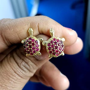 May include: A pair of gold-colored turtle stud earrings. The turtle's shell is covered in small, round, red gemstones. The earrings are displayed against a blue background.