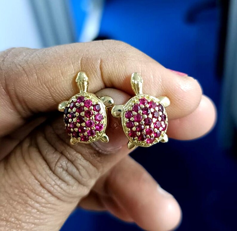 May include: A pair of gold-colored turtle stud earrings. The turtle's shell is covered in small, round, red gemstones. The earrings are displayed against a blue background.