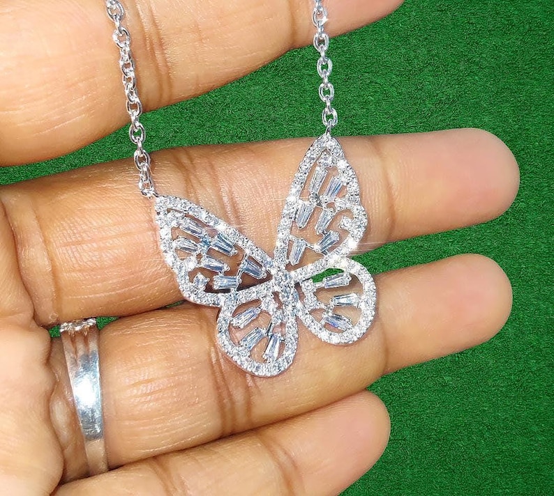 Peut inclure: Un collier pendentif papillon argent&eacute; avec une cha&icirc;ne d&eacute;licate. Le pendentif papillon est incrust&eacute; de pierres pr&eacute;cieuses claires et &eacute;tincelantes. Le collier est pr&eacute;sent&eacute; sur un fond vert.