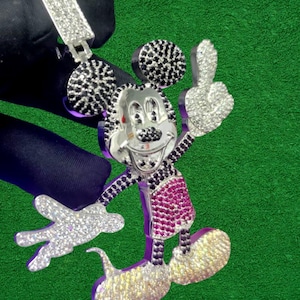 May include: A silver-toned pendant in the shape of Mickey Mouse, encrusted with sparkling white, black, and pink gemstones. The pendant features a bail for a chain and is set against a green background.