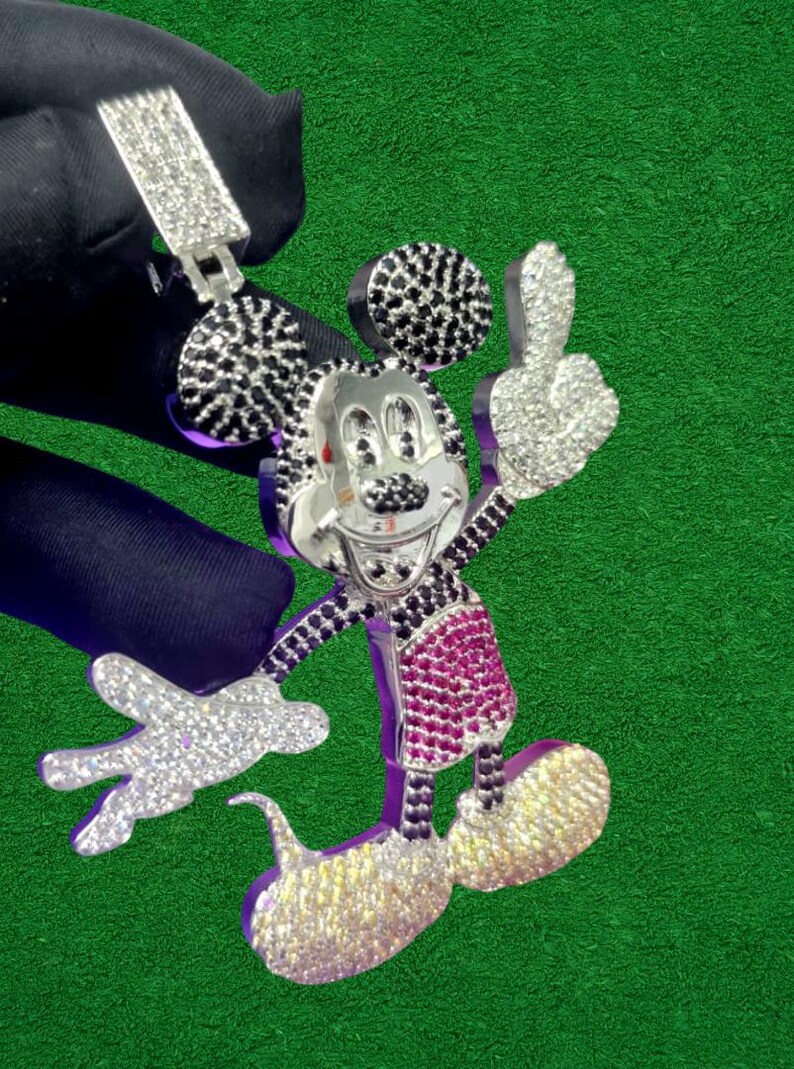 May include: A silver-toned pendant in the shape of Mickey Mouse, encrusted with sparkling white, black, and pink gemstones. The pendant features a bail for a chain and is set against a green background.