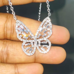 Peut inclure: Un collier pendentif papillon argent&eacute; avec une cha&icirc;ne d&eacute;licate. Le motif papillon est orn&eacute; de pierres pr&eacute;cieuses claires et scintillantes, cr&eacute;ant un effet &eacute;blouissant. Le collier est tenu dans une main sur un fond sombre.