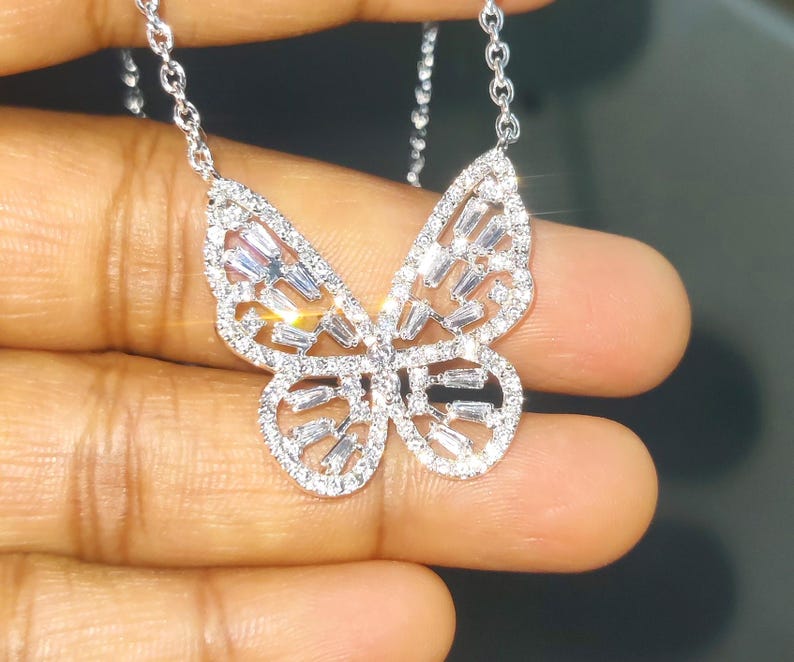 Peut inclure: Un collier pendentif papillon argent&eacute; avec une cha&icirc;ne d&eacute;licate. Le motif papillon est orn&eacute; de pierres pr&eacute;cieuses claires et scintillantes, cr&eacute;ant un effet &eacute;blouissant. Le collier est tenu dans une main sur un fond sombre.