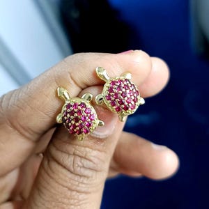 May include: A pair of gold-colored turtle stud earrings. Each turtle has a body covered in small, round, red gemstones. The earrings are displayed against a blurred background.