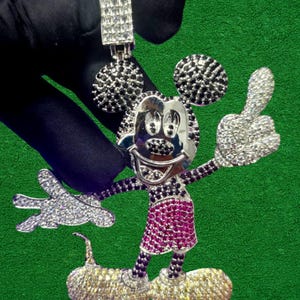 May include: A silver pendant in the shape of a cartoon mouse with a large head and round ears. The mouse has a smiling face, a red skirt, and yellow shoes. The pendant is covered in small, sparkling stones.
