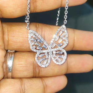Peut inclure: Un collier pendentif papillon argent&eacute;. Le motif papillon est orn&eacute; de petites pierres scintillantes, cr&eacute;ant un effet &eacute;blouissant. Le collier est tenu par une main, mettant en valeur les d&eacute;tails complexes du bijou.