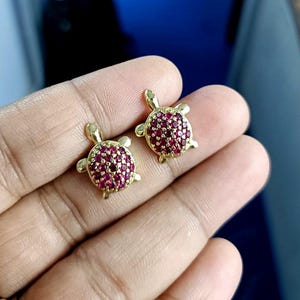 May include: A pair of gold-colored turtle-shaped earrings. The turtle shells are encrusted with small, round, red gemstones. The earrings are displayed against a blurred background.