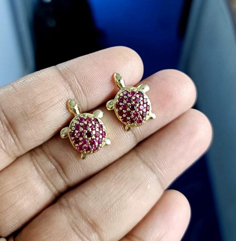 May include: A pair of gold-colored turtle-shaped earrings. The turtle shells are encrusted with small, round, red gemstones. The earrings are displayed against a blurred background.