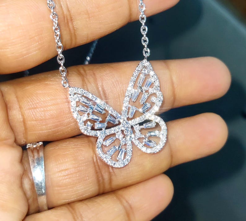 Peut inclure: Un collier pendentif papillon argent&eacute;. Le motif papillon est incrust&eacute; de petites pierres scintillantes. La cha&icirc;ne du collier est &eacute;galement argent&eacute;e et d&eacute;licate. Le bijou est pr&eacute;sent&eacute; sur un fond sombre.