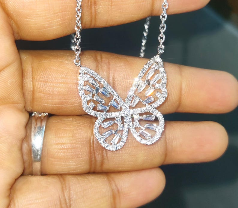 Peut inclure: Un collier pendentif papillon argent&eacute;. Le motif papillon est incrust&eacute; de petites pierres scintillantes, cr&eacute;ant un effet &eacute;blouissant. Le collier est tenu par une main, mettant en valeur les d&eacute;tails complexes du bijou. La cha&icirc;ne est d&eacute;licate et compl&egrave;te l'&eacute;l&eacute;gance du pendentif.