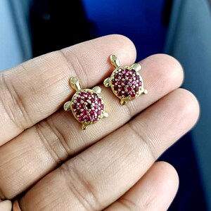 May include: A pair of gold-colored turtle stud earrings. The turtle shells are covered in small, round, red gemstones. The earrings are displayed against a blurred background.