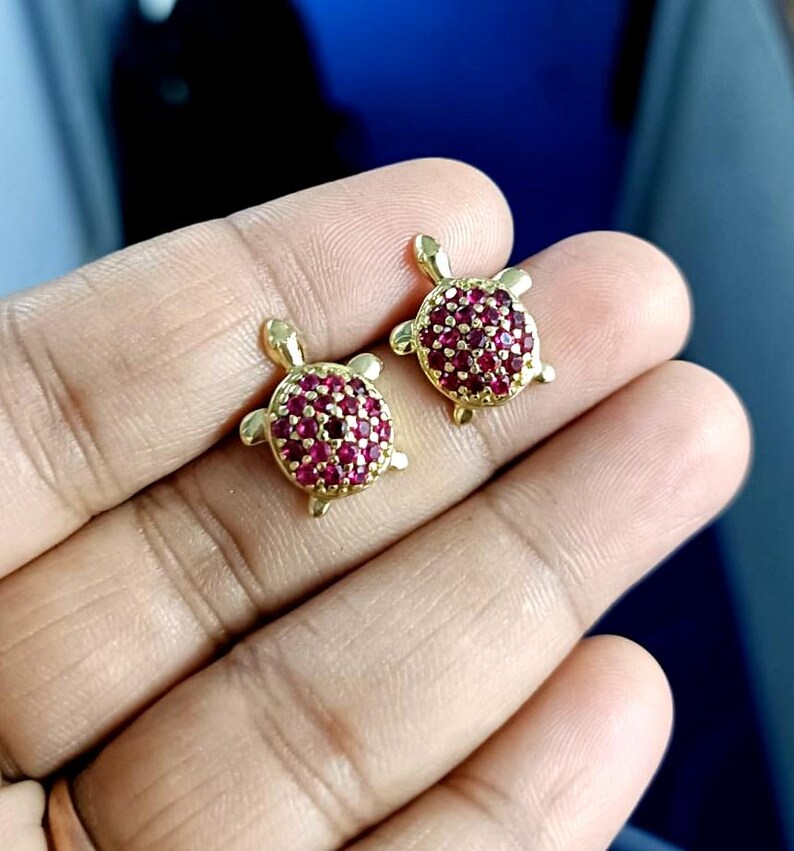 May include: A pair of gold-colored turtle stud earrings. The turtle shells are covered in small, round, red gemstones. The earrings are displayed against a blurred background.