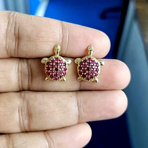 May include: A pair of gold-colored turtle stud earrings. The turtle's shell is covered in small, round, red gemstones. The earrings are displayed on a person's hand.