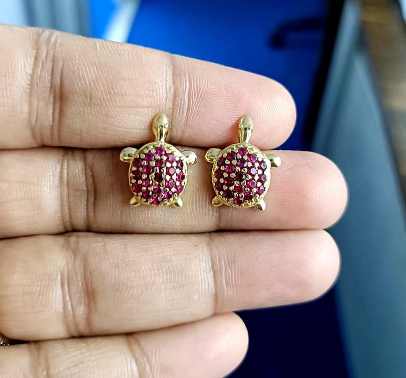 May include: A pair of gold-colored turtle stud earrings. The turtle's shell is covered in small, round, red gemstones. The earrings are displayed on a person's hand.