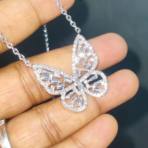 Peut inclure: Un collier pendentif papillon argent&eacute;. Le motif papillon est incrust&eacute; de petites pierres pr&eacute;cieuses transparentes. La cha&icirc;ne du collier est &eacute;galement argent&eacute;e et d&eacute;licate. Le pendentif est tenu dans une main.