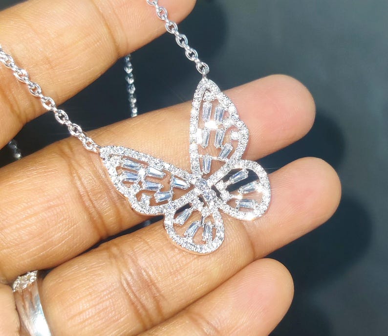 Peut inclure: Un collier pendentif papillon argent&eacute;. Le motif papillon est incrust&eacute; de petites pierres pr&eacute;cieuses transparentes. La cha&icirc;ne du collier est &eacute;galement argent&eacute;e et d&eacute;licate. Le pendentif est tenu dans une main.