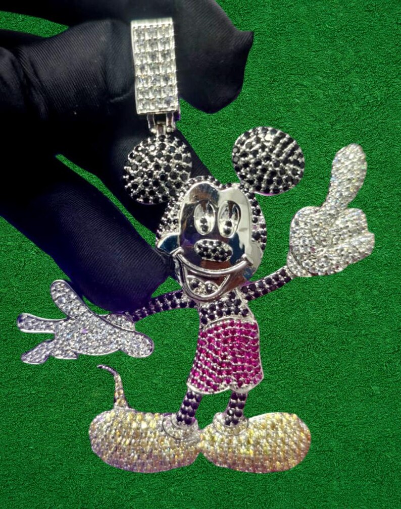 May include: A silver pendant in the shape of a cartoon mouse with a large head and round ears. The mouse has a smiling face, a red skirt, and yellow shoes. The pendant is covered in small, sparkling stones.