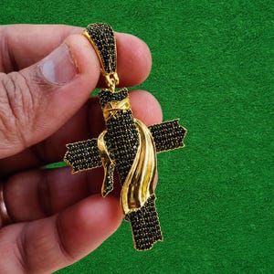 May include: Gold-toned cross pendant with black gemstone accents. The cross features a draped gold design and is held against a green background. The pendant is a religious accessory.