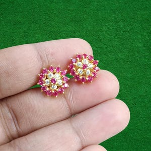 May include: A pair of gold-coloured stud earrings. Each earring has a floral design with a central cluster of white gemstones surrounded by a ring of red gemstones. The earrings are displayed against a green background.