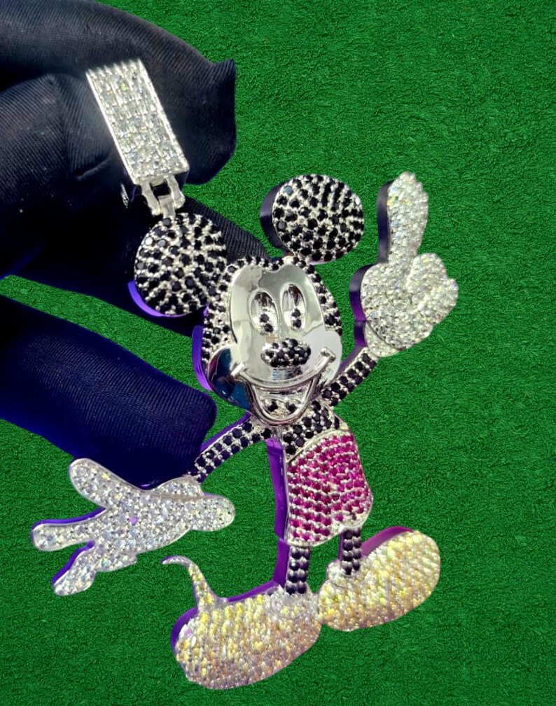 May include: A silver pendant featuring a cartoon character with black and white accents, a silver face, and a pink and white outfit. The pendant is encrusted with sparkling stones and is held up by a silver clasp.