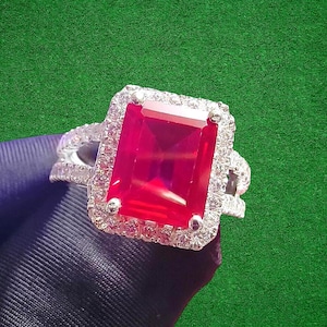 May include: A silver ring featuring a large, rectangular, vibrant red gemstone, surrounded by a halo of smaller, clear stones. The ring's band has a split design with additional small stones. The background is a solid green.