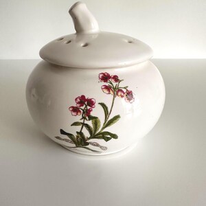Peut inclure: Un pot en céramique blanc avec un couvercle percé de trous et une petite poignée. Le pot est orné d'un motif floral rouge, rose et vert. Le pot rond est probablement utilisé pour le stockage ou la décoration. Un design classique.