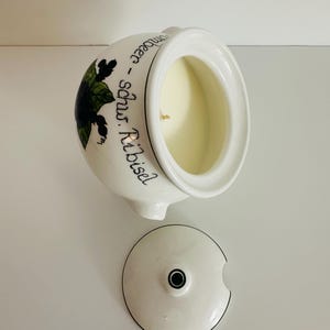 Puede incluir: Portavelas de cerámica blanca con tapa. El portavelas tiene una ilustración en negro y verde de una hoja y las palabras "schwarze Ribisel" en escritura negra. La tapa está quitada, revelando la vela en el interior.