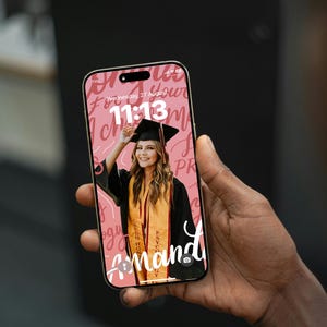 Puede incluir: Un teléfono inteligente que muestra un fondo de pantalla con temática de graduación. La pantalla muestra una foto de una graduada con birrete y toga, con el nombre "Amanda" y la hora "11:13". El fondo es rosa con texto en escritura.