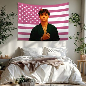 Puede incluir: Un tapiz con un retrato de una persona frente a un diseño de bandera estadounidense rosa y blanco. La persona lleva una chaqueta verde oscuro y collares dorados. El tapiz cuelga sobre una cama con ropa de cama blanca y una manta marrón.