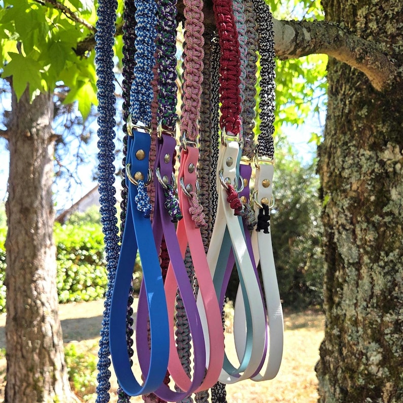 May include: A collection of dog leashes featuring braided paracord handles in various colors including blue, purple, pink, and white. Each leash includes a smooth, solid-colored silicone loop handle attached with metal rivets and a metal ring.
