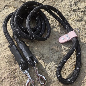 Puede incluir: Una correa para perro de cuerda negra con detalles plateados reflectantes tejidos en el cordón. La correa incluye dos ganchos giratorios de metal plateado, tubos termorretráctiles negros y una pieza decorativa de color rosa claro.