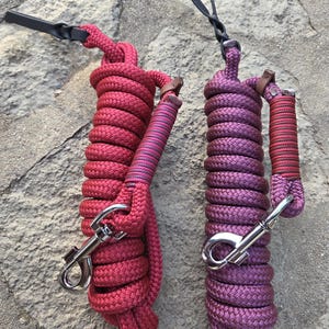 Könnte beinhalten: Zwei aufgerollte Führstricke mit silberfarbenen Metallkarabinerhaken. Ein Strick ist in einem einheitlichen Dunkelrot gehalten, der andere in einer violett-roten Mischung. Beide Stricke haben eine geflochtene Struktur, einen Lederakzent und einen schwarzen Riemen.